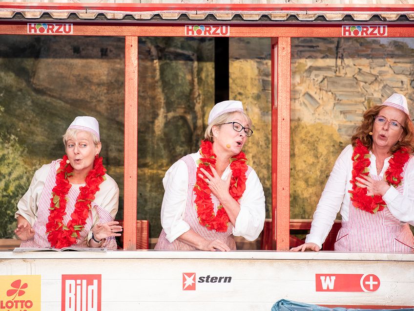 Drei Frauen singen in einem Bühnen-Kiosk. Sie tragen Imbiss-Kleidung und rote Girlanden.