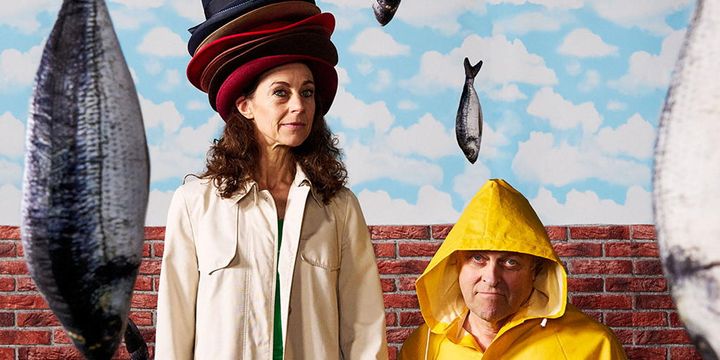eine Frau und ein Mann im Regenmantel stehen vor einer Mauer aus Ziegelsteinen, rundherum hängen Fische in der Luft