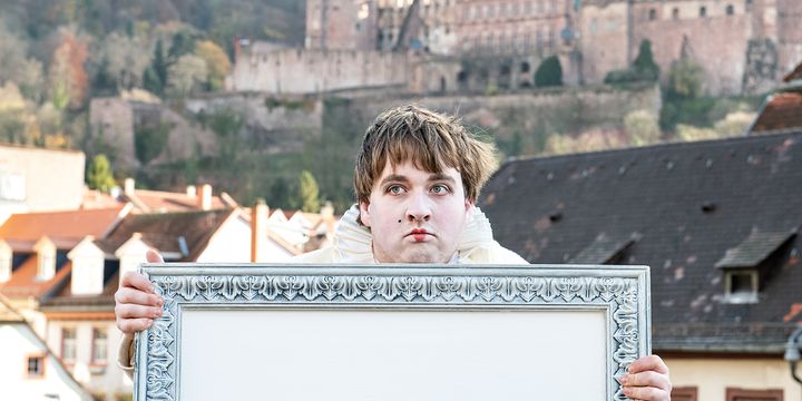 Der Schauspieler hält einen leeren Bilderrahmen in der Hand, im Hintergrund ist das Heidelberger Schloss zu sehen