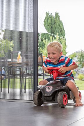 Kind mit Bobby-Car fährt durch Terrassentür mit Insektenschutz