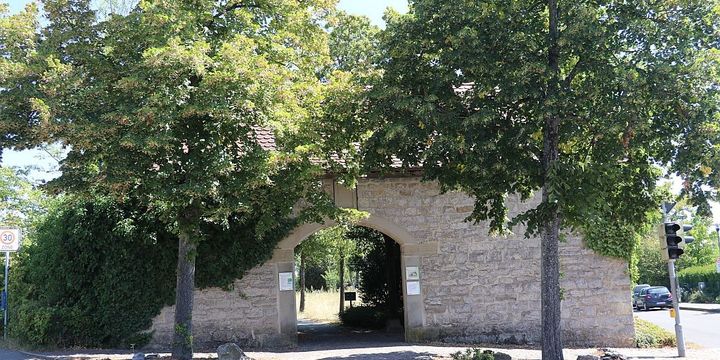 Hier ist das steinerne Torhaus des Friedhof Rutesheim zu sehen, mit den beiden grünen Laubbäumen rechts und links davor.