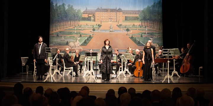 Die Sänger*innen Rupert Enticknap, Franziska Gottwald und Gaëlle Mechal mit dem Orchester, im Hintergrund ein Bild des Schlossgartens Schwetzingen